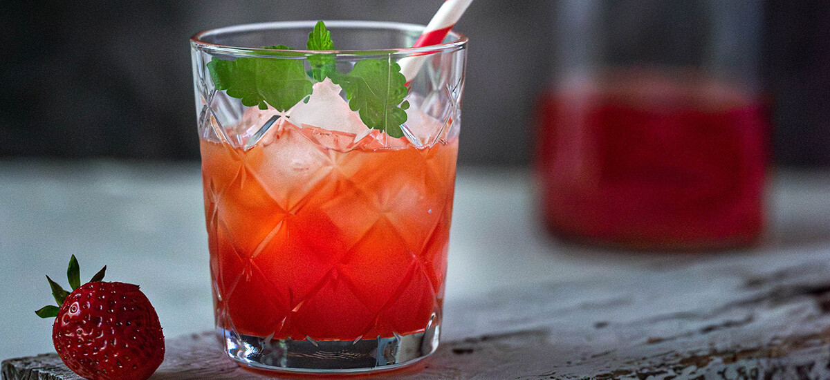 Ein roter Drink mit Eiswürfeln und Strohhalm steht auf einem Holzbrett und daneben liegt eine Erdbeere